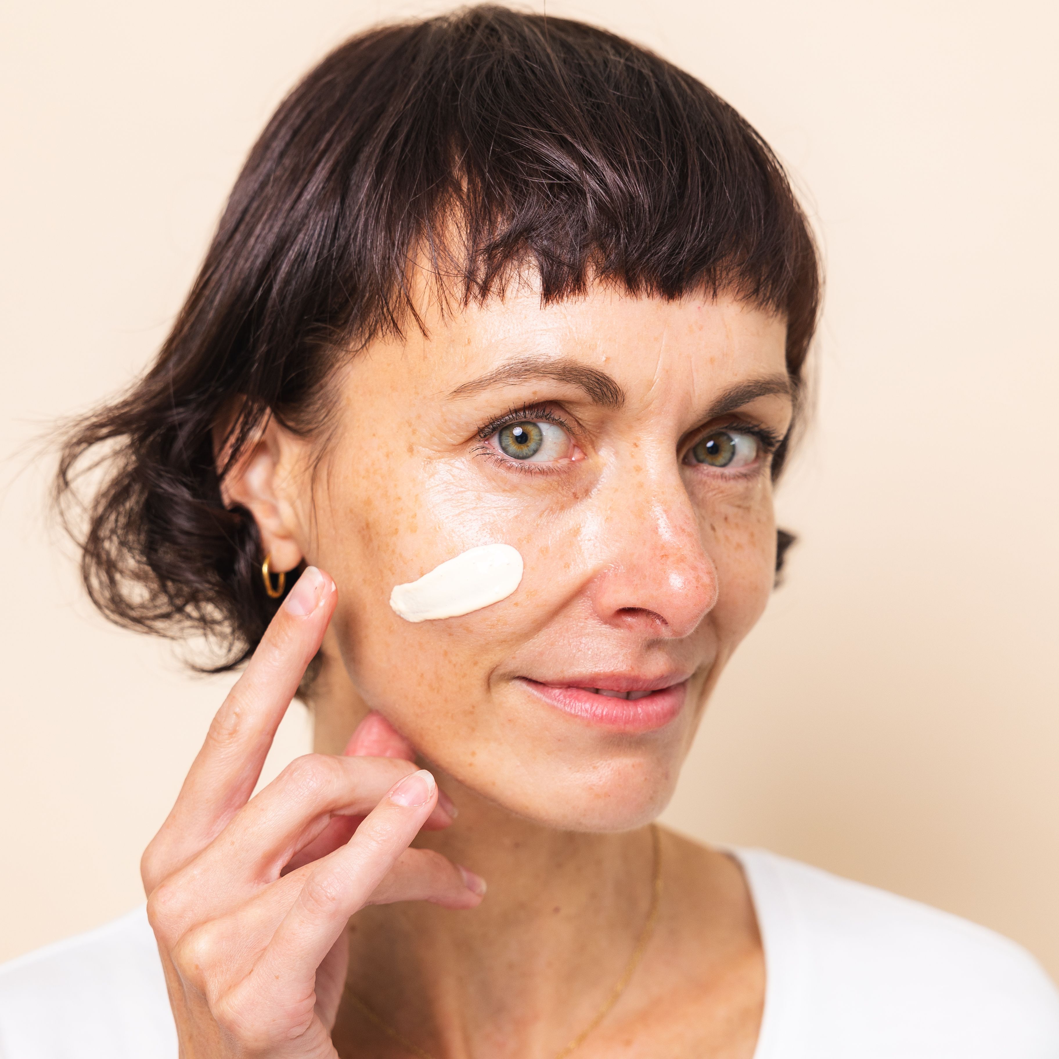 Natürliche Frau Anfang 40 mit kurzem, dunkelbraunen Haar blickt in die Kamera und trägt sich die Hautcreme+ von Beyer & Söhne aufs Gesicht auf